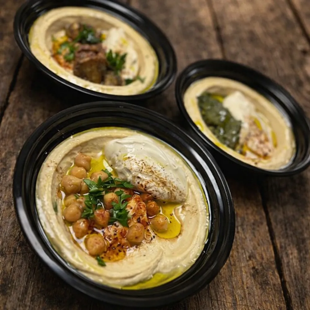 Three Hummus Y'all bowls on a wooden table, the front bowl topped with chickpeas, tahini, parsley and olive oil
